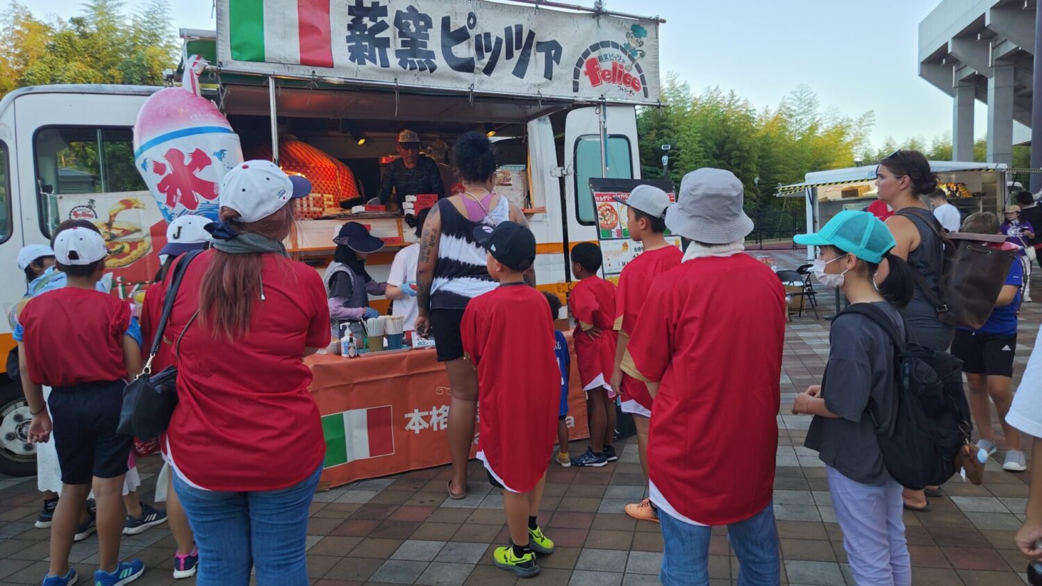 日米対抗ソフトボール2023第1戦の会場となった山口県岩国市の絆スタジアム（愛宕スポーツコンプレックス）に出店されたピザ店に集まる日米のソフトボールファンたち-Journal-ONE撮影