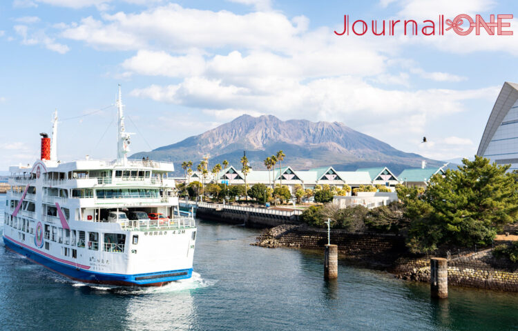 雄大な桜島を背景に鹿児島港を出港する「第二桜島丸」。白地にピンクとブルーのラインが入った船体が、青い海と空に映える美しい風景。