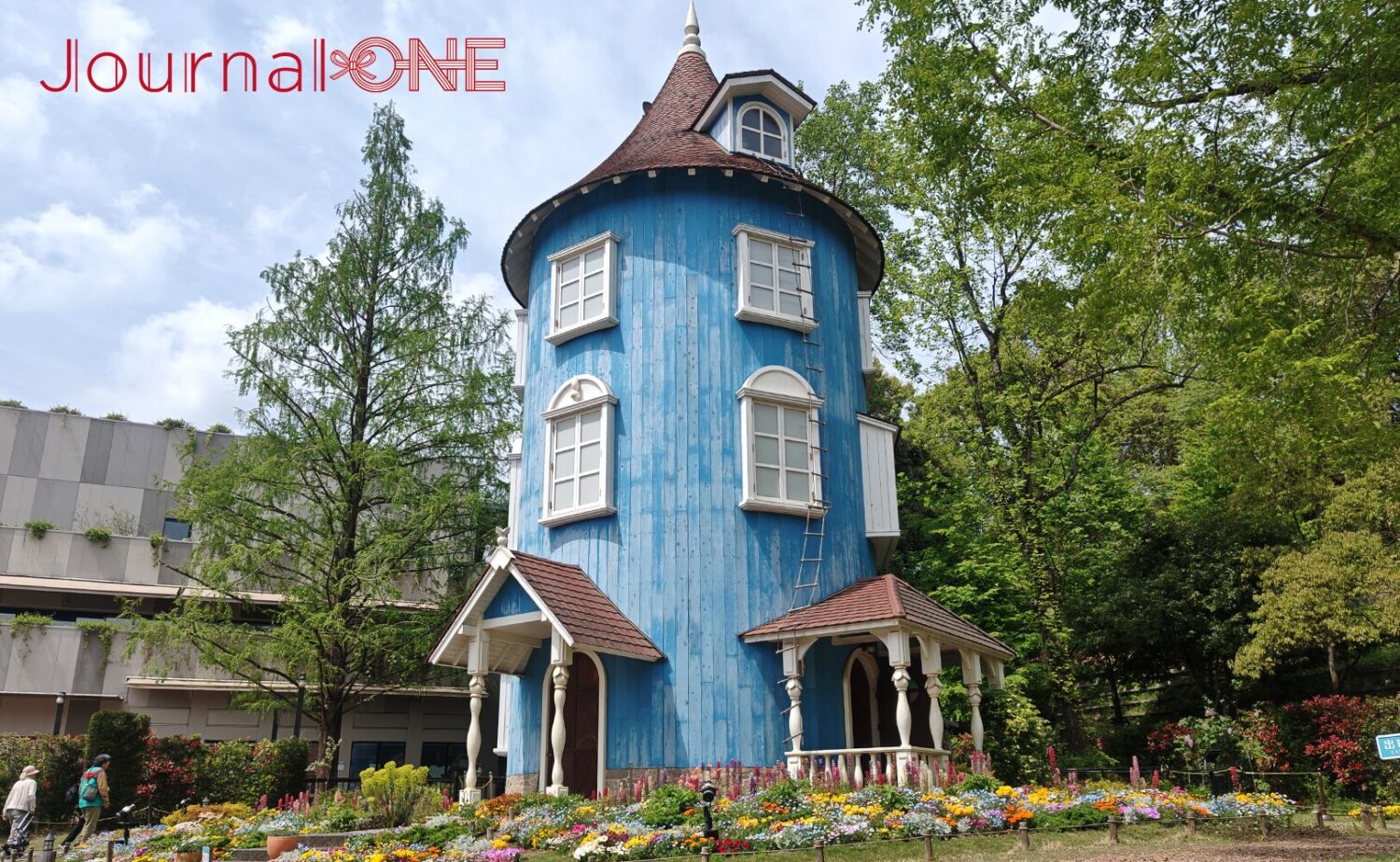 ムーミンバレーパークのシンボル「ムーミン屋敷」。春の花々が咲く庭園の奥に、青い板張りの壁と赤い屋根の円筒形の建物が建っている。周囲には豊かな木々。