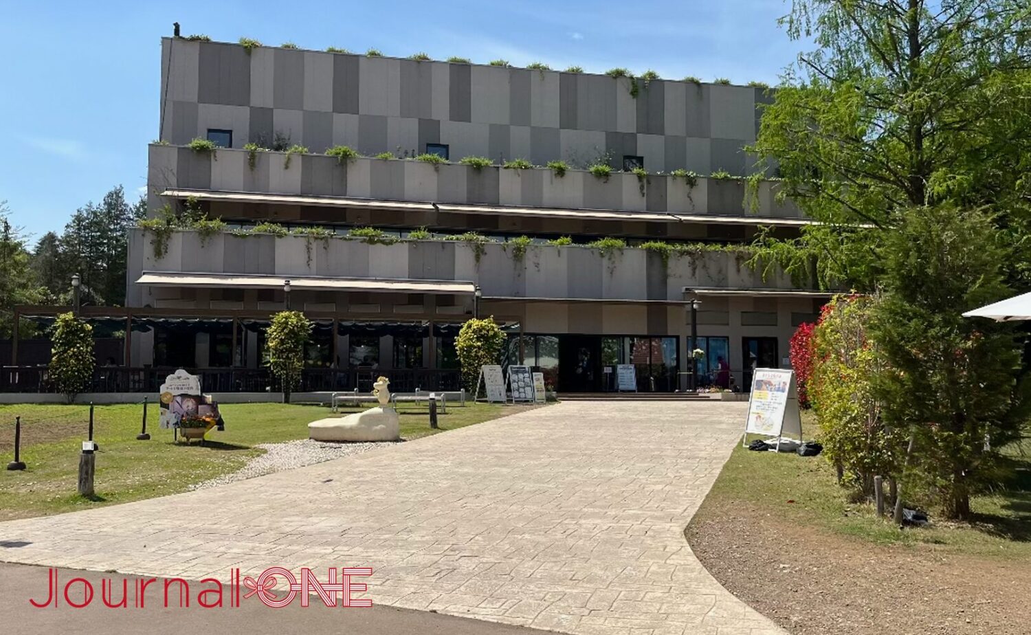ムーミンバレーパーク内の地上3階建て展示施設「コケムス（KOKEMUS）」。グレーのモダンな外壁にテラス席が並び、手前には芝生と石畳の通路、そしてニョロニョロのオブジェが置かれた開放的な外観。