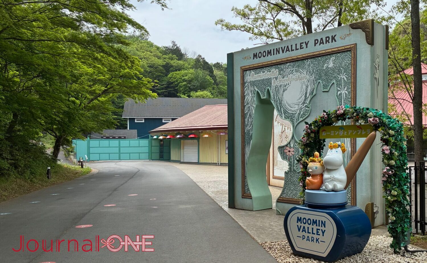 ムーミンバレーパークのエントランス。ムーミンの物語を模した巨大な本のオブジェがゲートとなり、その先には新緑の並木道が続く。「ムーミン谷とアンブレラ」の装飾を施したムーミンとリトルミイのフォトスポットが並ぶ様子。