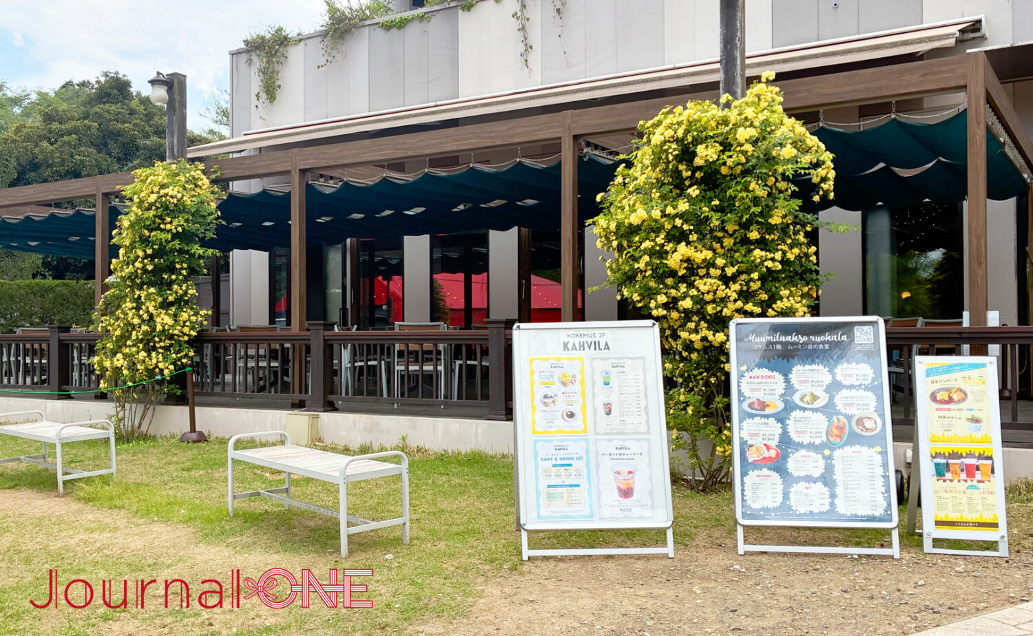 埼玉県飯能市のムーミンバレーパーク内、コケムスの「ムーミン谷の食堂」テラス席。柱に絡まる黄色のモッコウバラが満開を迎え、紺色のシェードが設置された開放的な屋外スペース。手前にはメニュー看板が並んでいる。