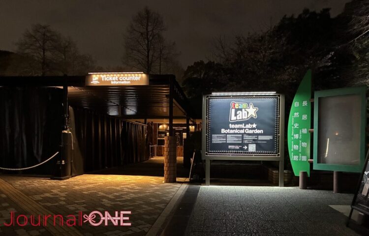 長居植物園の深い夜の静寂に浮かび上がる、チームラボ ボタニカルガーデン 大阪の入場ゲート。漆黒の闇に包まれたチケットカウンターへの導線と、それを照らす「TeamLab ★ Botanical Garden」の鮮明な光のロゴ看板。人工的な光と周囲の自然（木々）が交差する、知性的な夜の入口。