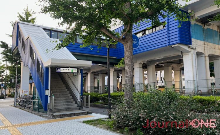 大阪メトロ(Osaka Metro)中央線の「大阪港駅」の地上出入口周辺の景観。歩道に面して設置されたコンクリート造の階段の横に、鮮やかなブルーの波板状の外壁と白い屋根で覆われた通路構造が伸び、頭上には高架駅へと接続する同じくブルーの駅舎の一部が頑強な白い橋脚に支えられている。
