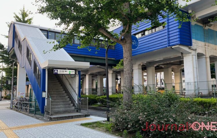 大阪メトロ（Osaka Metro）中央線の「大阪港駅」の地上出入口周辺の景観。歩道に面して設置されたコンクリート造の階段の横に、鮮やかなブルーの波板状の外壁と白い屋根で覆われた通路構造が伸び、頭上には高架駅へと接続する同じくブルーの駅舎の一部が頑強な白い橋脚に支えられている。