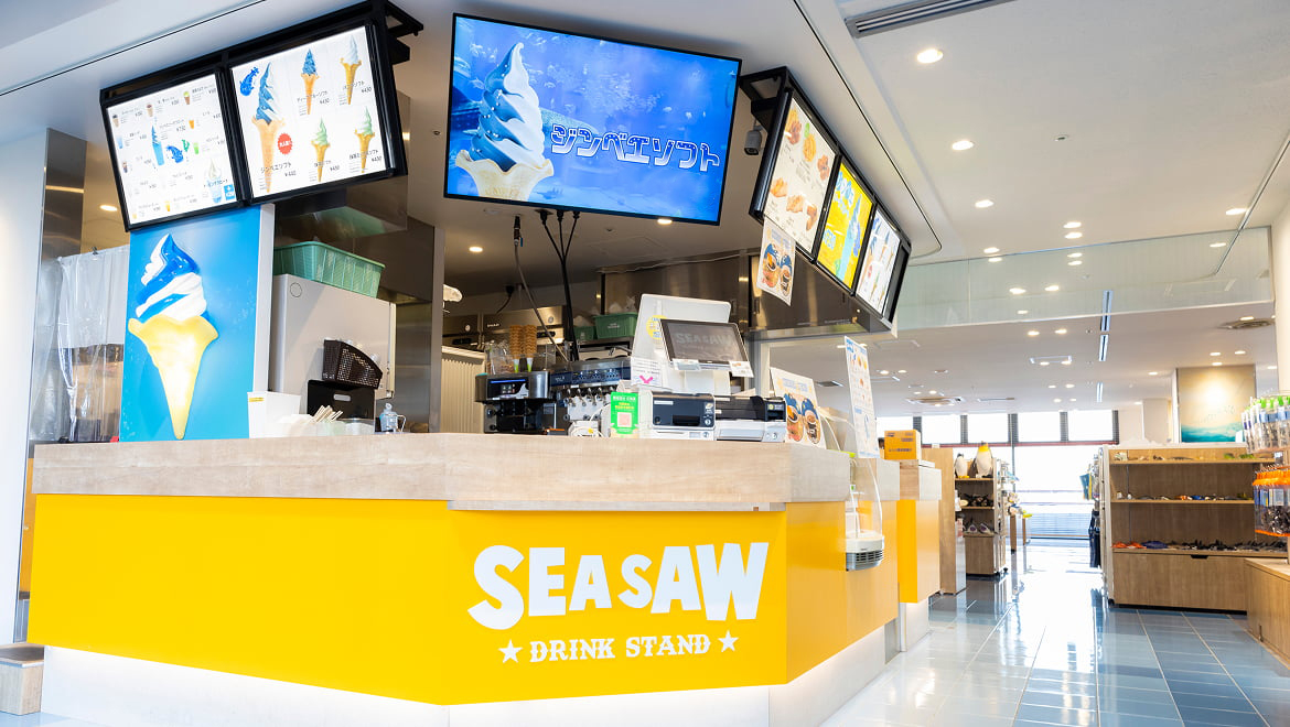 大阪港エリアの水族館「海遊館」の出口改札外にある、無料ゾーンのドリンクスタンド「SEA SAW（シーソー）」。白を基調とした明るい店内に、鮮やかな黄色のカウンターが配され、正面には「SEA SAW DRINK STAND」の白いロゴが描かれている状態。カウンター上部には液晶モニターが設置され、水色と白のストライプが施された名物「ジンベエソフト」のイメージ画像が映し出されている。