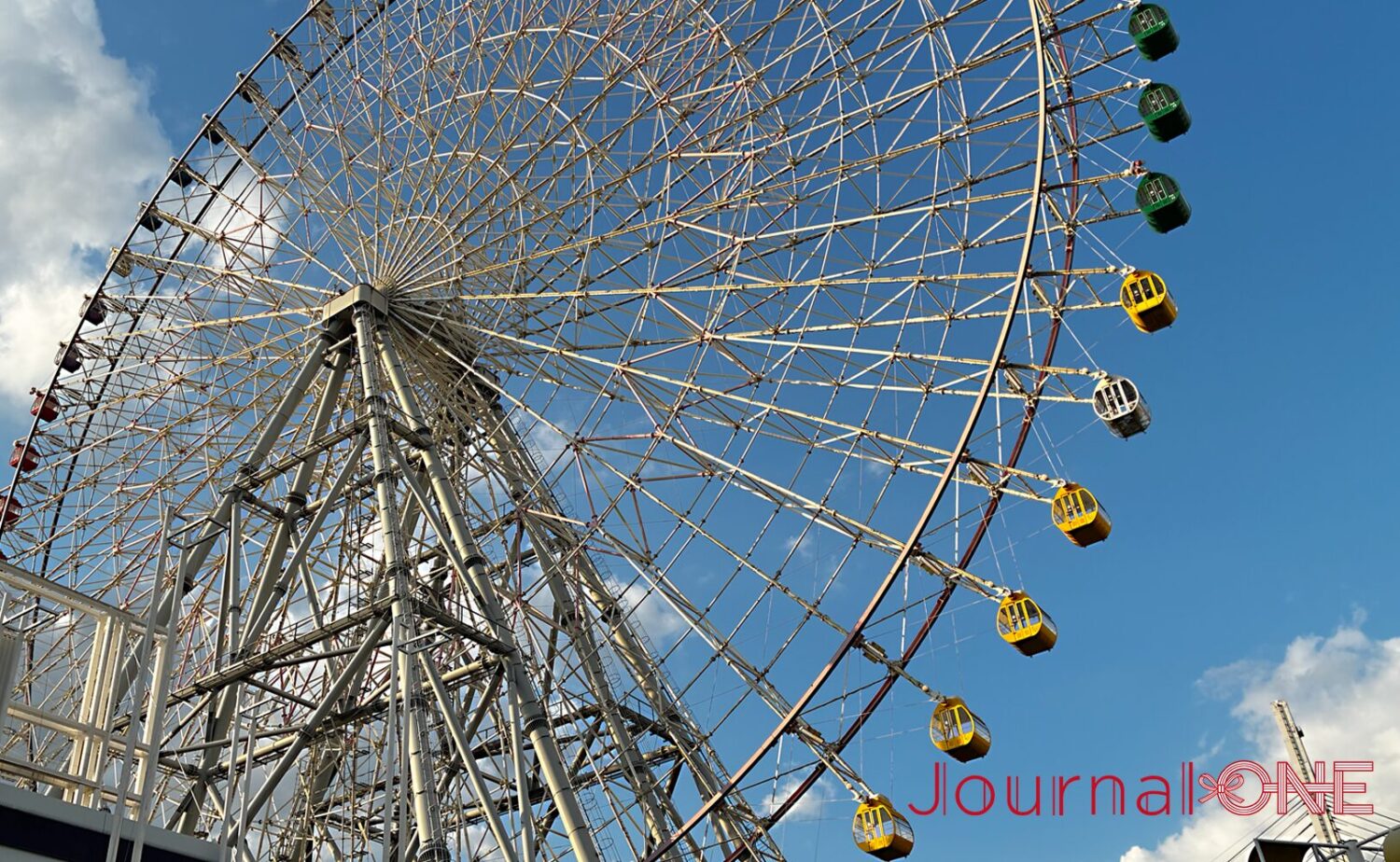 青空を背景に斜め下から見上げた天保山大観覧車。緻密に組まれた白い鉄骨トラス構造の円周に沿って、緑や黄色、白のカラフルなゴンドラが規則的に並んでいる状態。約15分間のゆるやかな回転で大阪湾や市街地のパノラマ景観を提供。