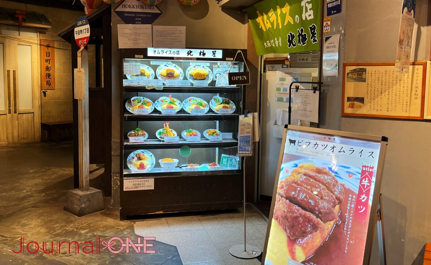 天保山マーケットプレイス内の「なにわ食いしんぼ横丁」に出店している、オムライス発祥の老舗洋食店「北極星 天保山店」の店舗前。昭和30年代の街並みを再現した薄暗い通路沿いに、様々な種類のオムライスの食品サンプルが明るくライトアップされた4段のショーケースが設置され、その右上には「オムライスの店 北極星」と染め抜かれた緑色の暖簾が掛けられている。
