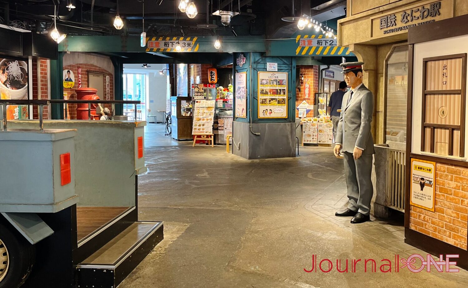 大阪市港区の天保山マーケットプレース内にあるフードテーマパーク「なにわ食いしんぼ横丁」のレトロな街並み。右手前には「国鉄 なにわ駅」の看板や出札口の意匠と共に、制服と制帽を着用した駅員の等身大人形が佇み、左側には当時の乗り物を模した造作物のステップが見える状態。奥へと続く通路には、レトロな電球照明や「注意 桁下2.3米」の標識が施された高架下の梁、赤い丸ポストなどが精緻に再現されている。