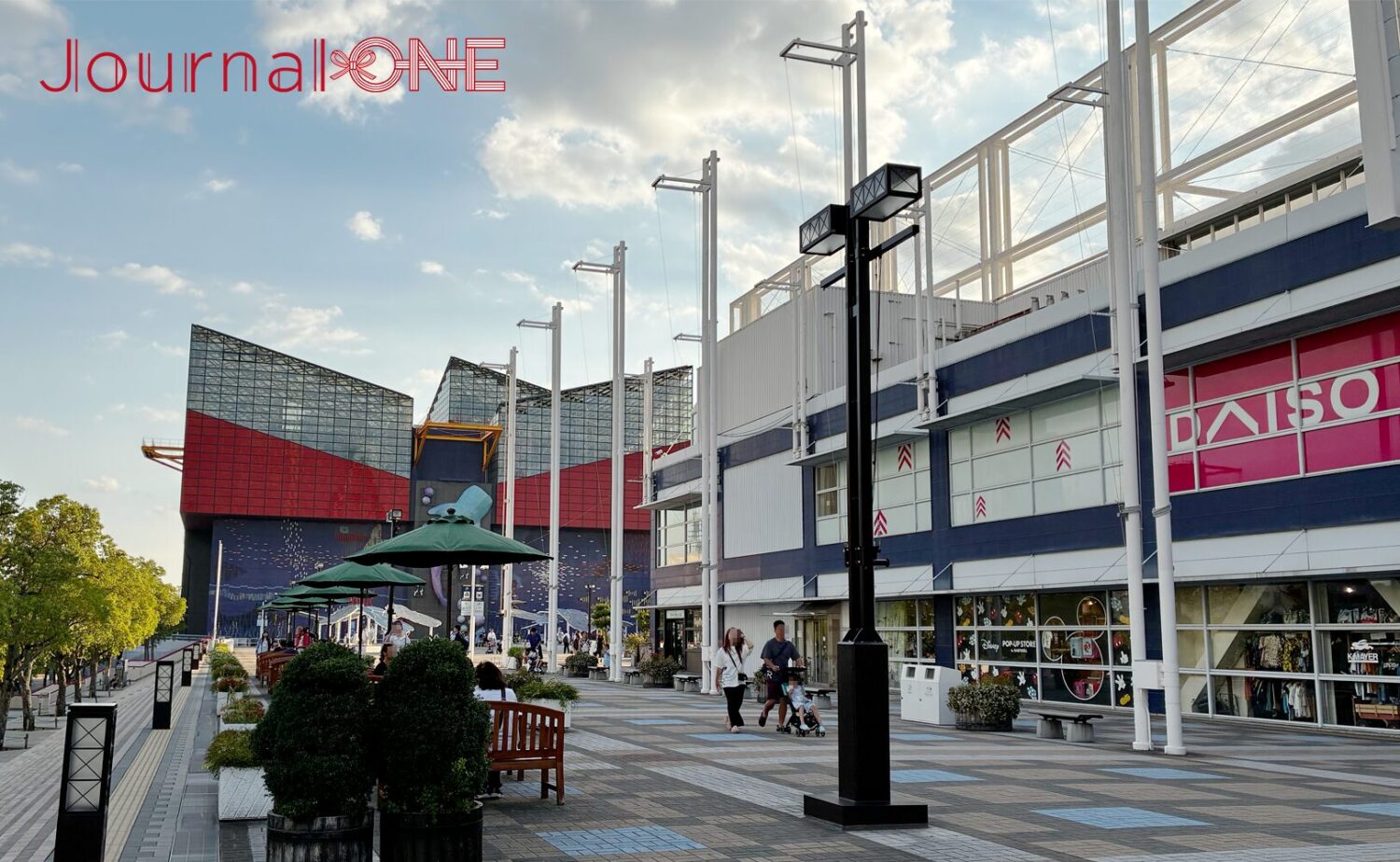 大阪港の複合商業施設「天保山マーケットプレイス」に隣接する広場。手前にはインターロッキングが敷かれた遊歩道に沿ってパラソル付きのテラス席や植栽プランター、近代的なデザインの街灯ポールが整然と並び、右側には商業施設の建物外観が、奥には赤と青の特徴的な外壁と傾斜したガラス屋根を持つ水族館「海遊館」の巨大な建築がそびえている。