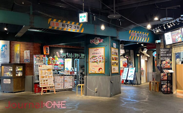 天保山マーケットプレイス内にあるフードテーマパーク「なにわ食いしんぼ横丁」の一角。高架下の意匠を再現した「注意 桁下2.3米」などの標識が施された鉄骨風の梁が頭上を通り、中央の太い柱には「なにわ食いしんぼ横丁 会場内案内マップ」の看板が設置されている。