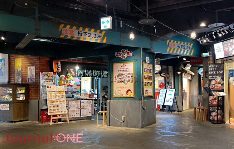 天保山マーケットプレイス内にあるフードテーマパーク「なにわ食いしんぼ横丁」の一角。高架下の意匠を再現した「注意 桁下2.3米」などの標識が施された鉄骨風の梁が頭上を通り、中央の太い柱には「なにわ食いしんぼ横丁 会場内案内マップ」の看板が設置されている。