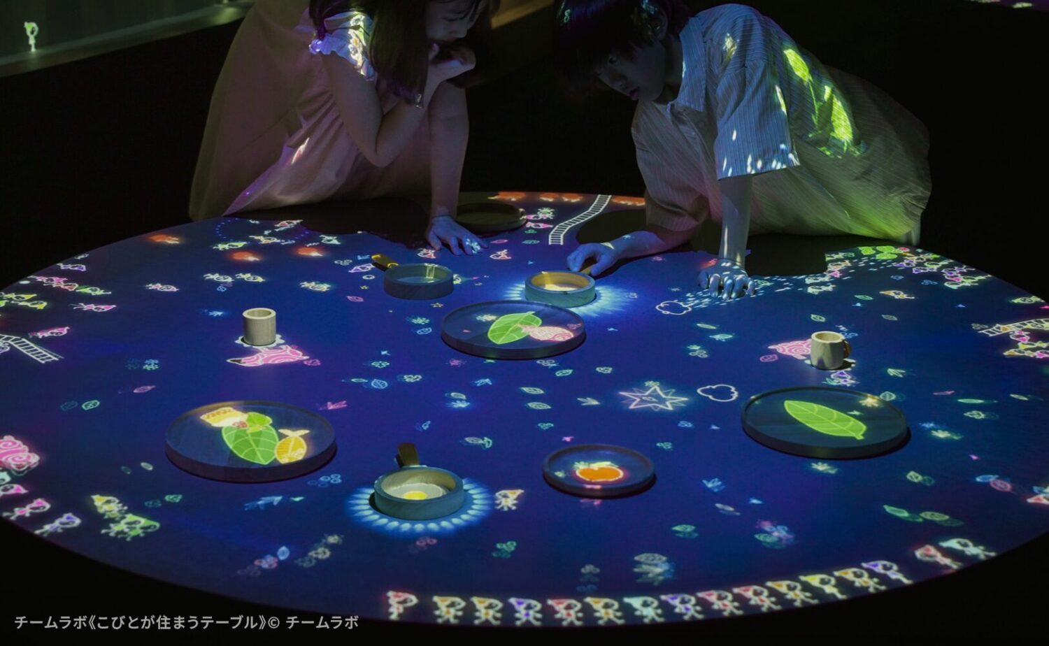 teamLab Biovortex KYOTO（チームラボバイオヴォルテクス京都）」内で展示されているチームラボのアート作品《こびとが住まうテーブル》。円形のテーブルの上にフライパンや皿など様なオブジェクトが置かれ、その上を動き回る小さな「こびと」やグラフィックの光が、鑑賞者が置いた手や物に反応して飛び跳ねたり走ったりするインタラクティブな作品空間。