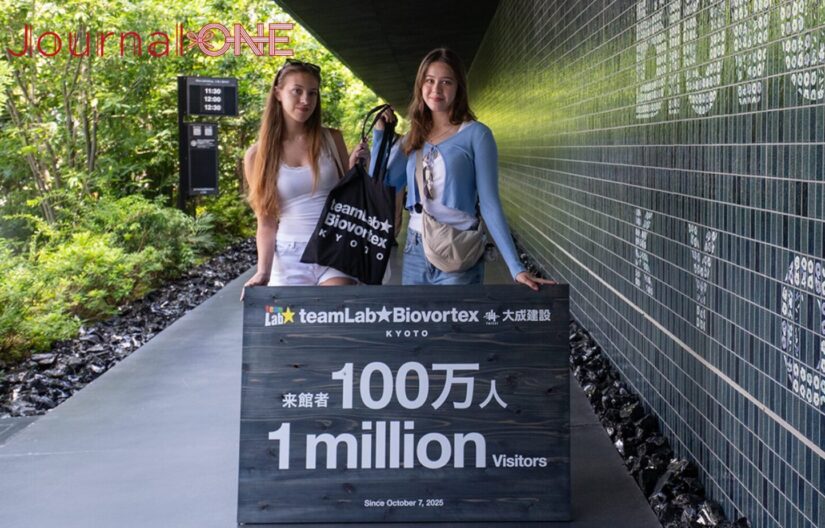 「teamLab Biovortex KYOTO（チームラボバイオヴォルテクス京都）」のアプローチ空間で行われた、来館者100万人達成の記者会見。手前に置かれた「来館者 100万人 1 million Visitors」と書かれた木製の記念看板を挟み、記念のオリジナルトートバッグを手にする2人の外国人女性来館者。右側に続く深い緑色のタイル壁面と、左側に広がる豊かな植栽の緑に囲まれた通路で、歴史的な節目を祝うセレモニーの様子を捉えた。