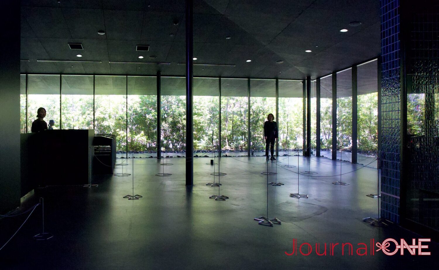 「teamLab Biovortex KYOTO(チームラボバイオヴォルテクス京都)」の1階エントランスロビー。背後に一面の緑豊かな庭園が広がる大きな窓があり、手前には鉄道の改札口を思わせる黒い入場ゲートが、来客を待つ。