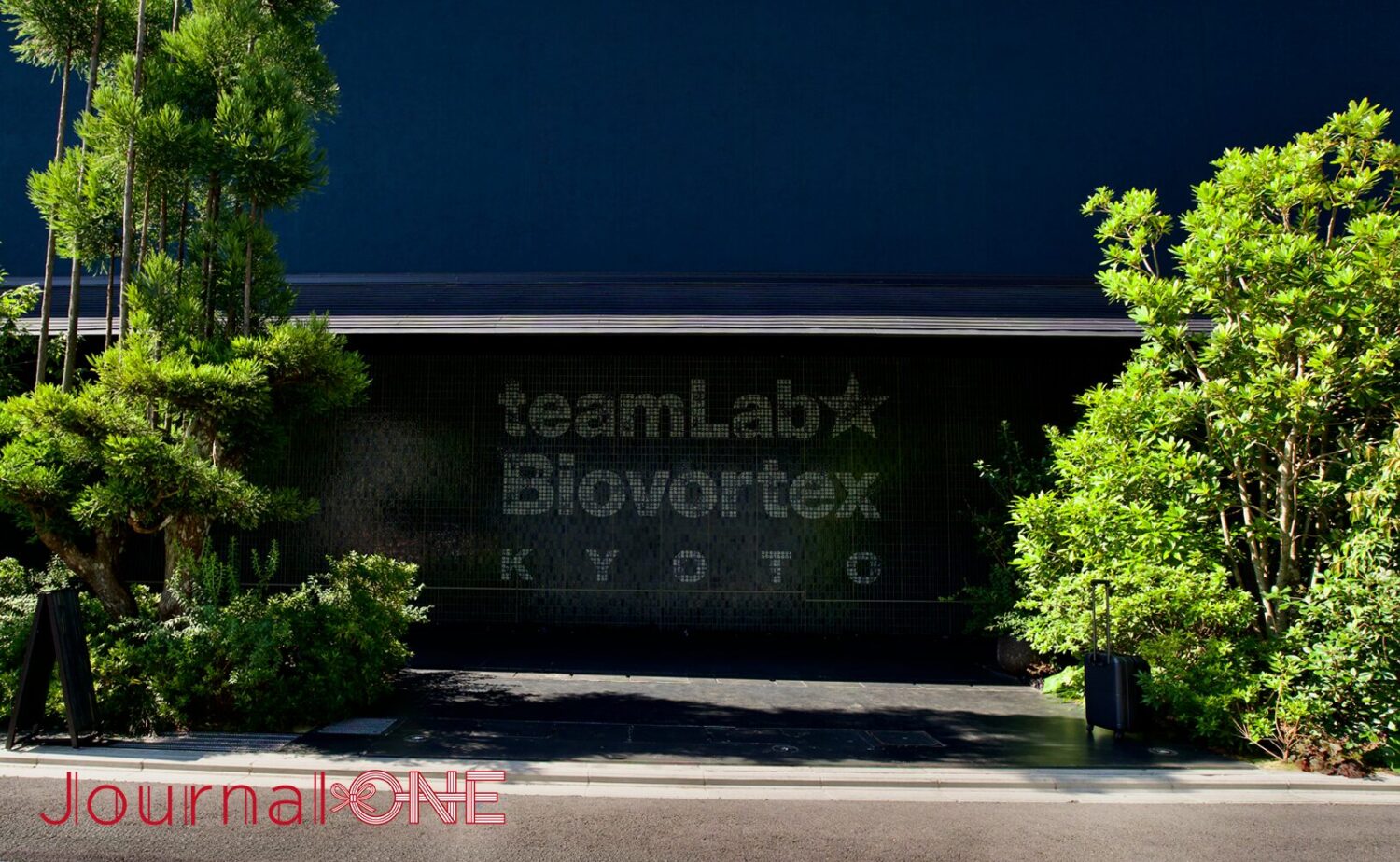 京都に位置する「teamLab Biovortex KYOTO(チームラボバイオヴォルテクス京都)」のファサード。黒いタイル壁面の中央に浮かび上がる「teamLab Biovortex KYOTO」の白いロゴマークと、その左右を囲むように配置された青々とした松や植栽の緑、そして上部に広がる深い紺色の外壁が織りなす、モダンと伝統が融合したミュージアムエントランスの落ち着いた佇まい。