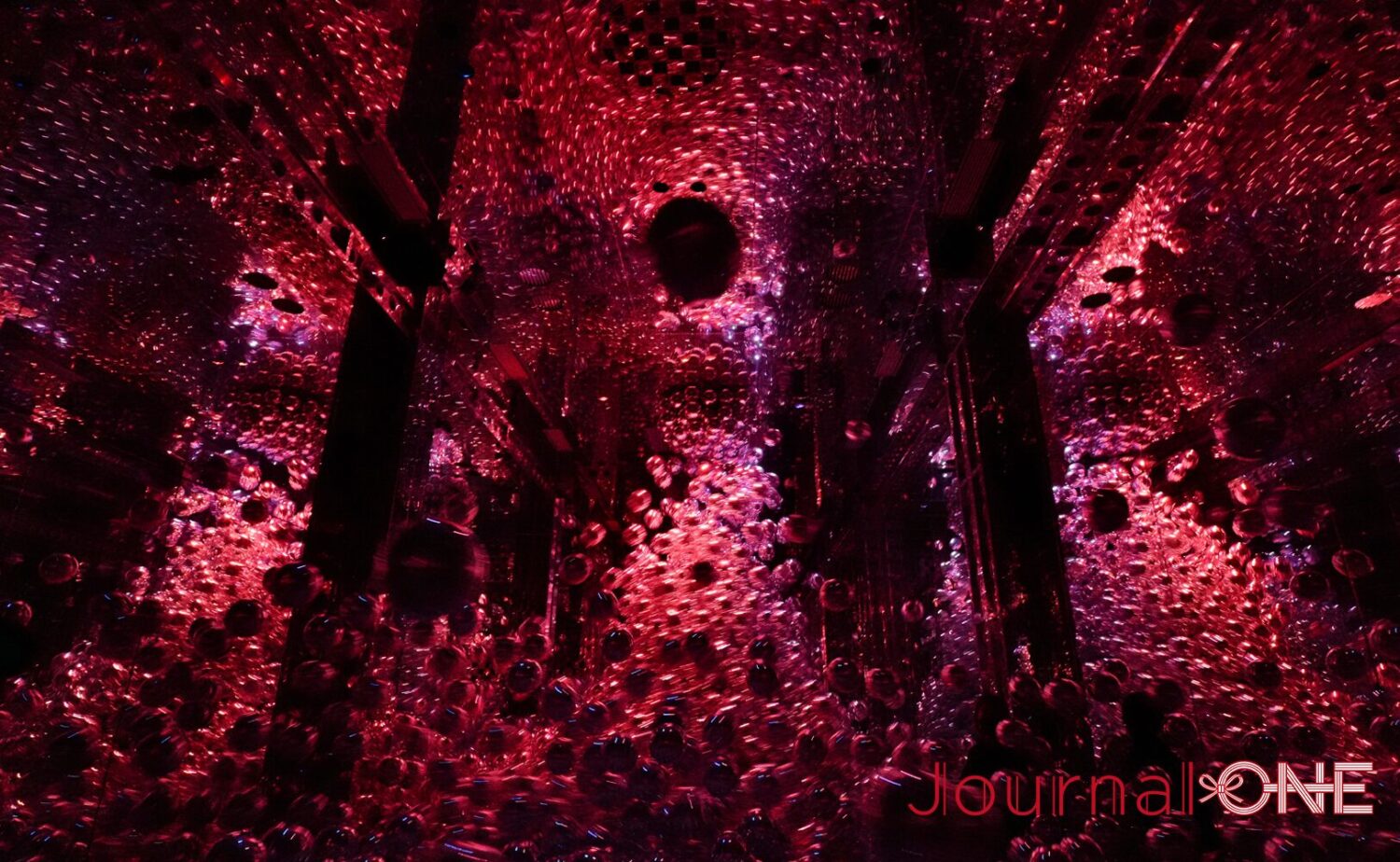 teamLab Biovortex KYOTO(チームラボバイオヴォルテクス京都)」内で展示されているチームラボのアート作品《変容する連続体》。暗闇に包まれた鏡面の展示空間全体に、無数の球状の光や赤・紫調の光の粒子が幾何学的かつ高密度に波及・浮遊し、無限に広がる光の粒子群と黒い構造フレームが交錯する幻想的な作品空間。