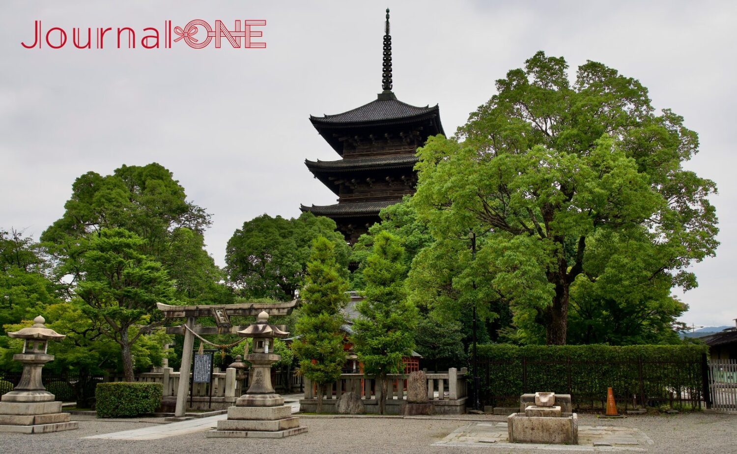 京都の南区九条町にある東寺(教王護国寺)の象徴である国宝・五重塔。手前に石灯籠や鳥居、注連縄が配された敷地があり、周囲を取り囲む豊かな落葉樹や生垣の緑の奥に、重厚な本瓦葺きの木造五層の塔がそびえ立つ、曇り空の下の厳かな歴史的景観。