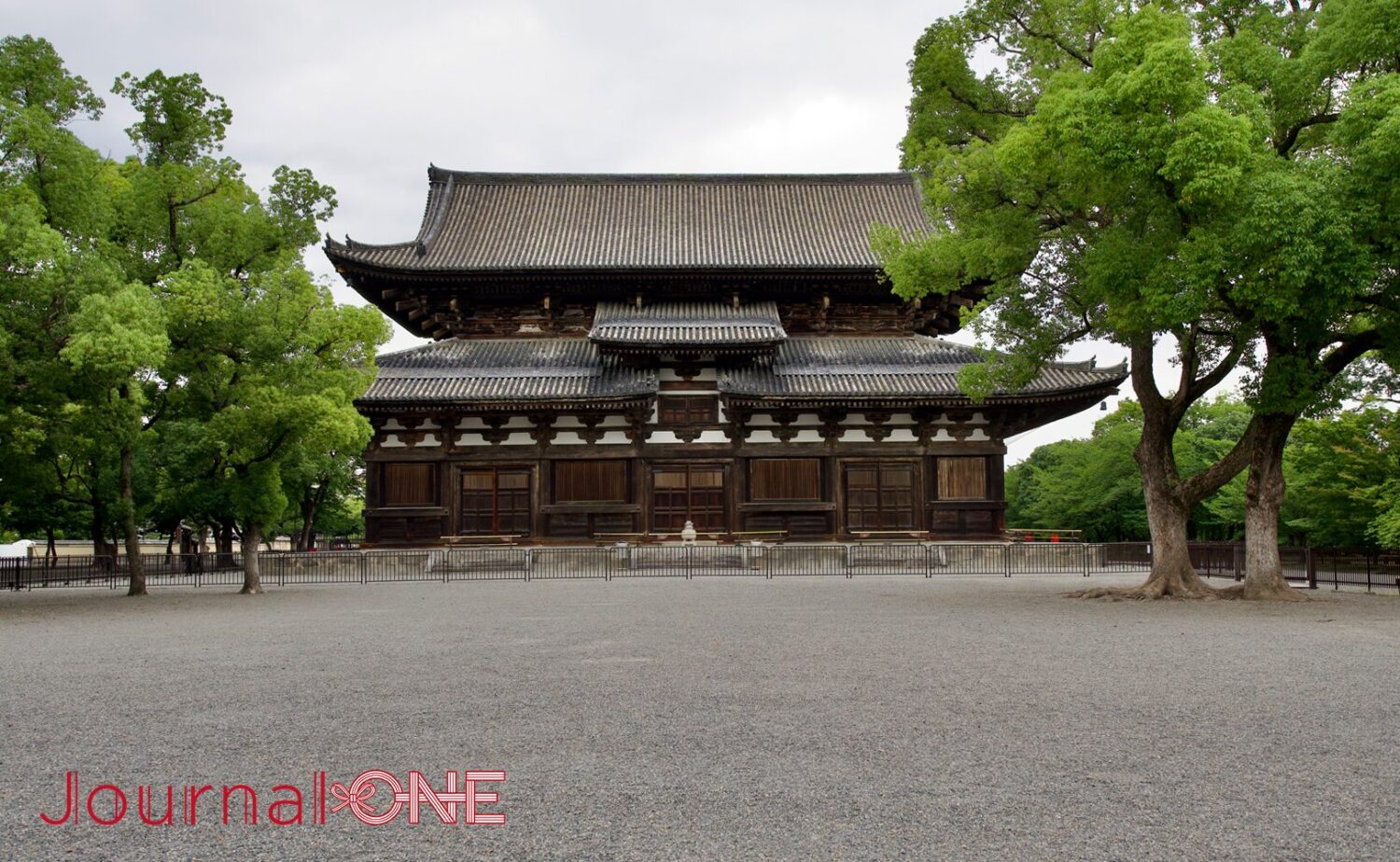 京都の南区九条町に位置する東寺(教王護国寺)の重要文化財である講堂。手前に広がる砂利敷きの平地と、左右にそびえ立つ大きな落葉樹の緑に囲まれるようにして、重厚な瓦屋根と漆喰壁、木造構造が美しい入母屋造の仏堂が静かに佇む、曇り空の下の厳かなシチュエーション。