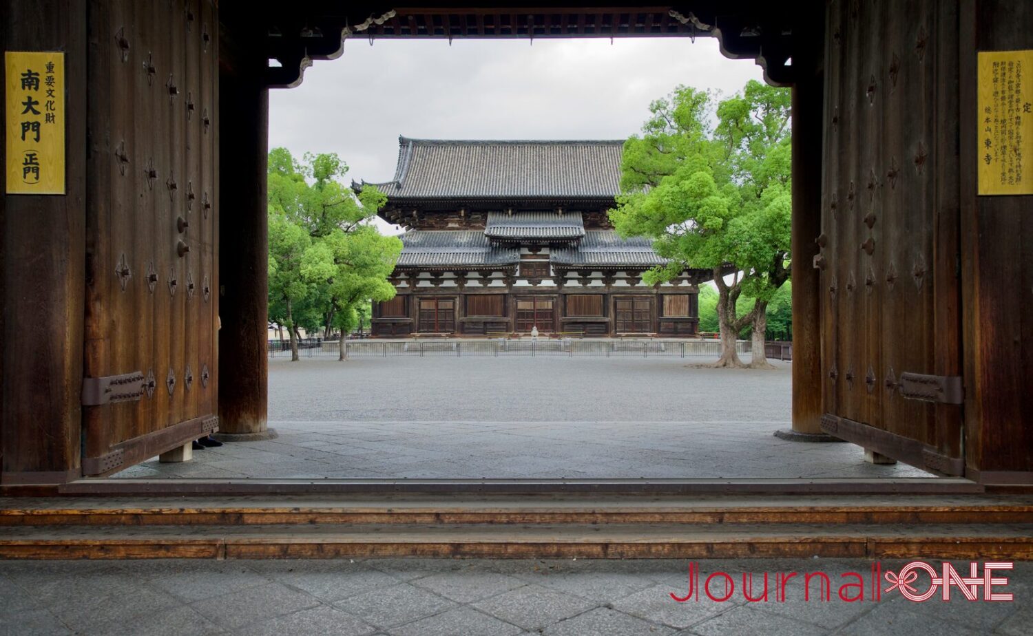 京都の南区九条町にある東寺（教王護国寺）の重要文化財・南大門（正門）。大きく開かれた木製の頑丈な門扉と柱のフレーム越しに、砂利敷きの境内の中央に佇む入母屋造の講堂と周囲を囲む新緑の落葉樹、そして曇り空の落ち着いた光に包まれた厳かな歴史的景観。